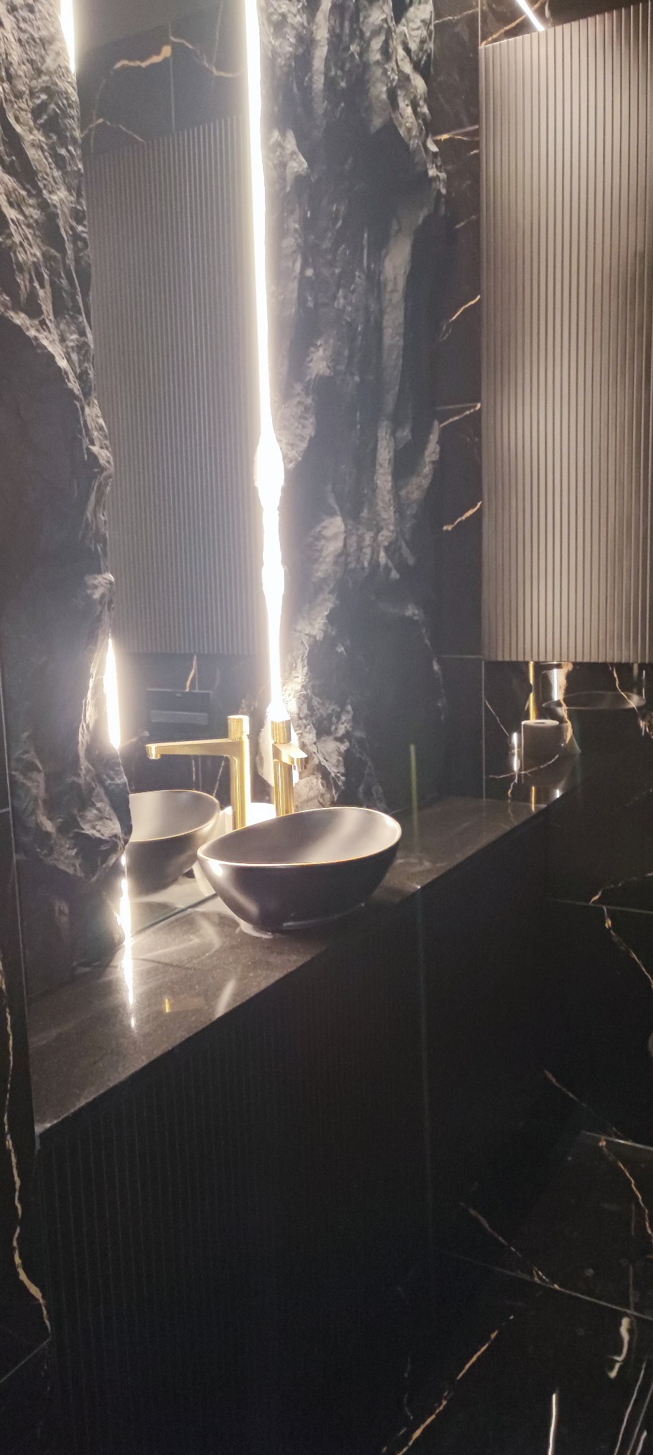 Nowoczesna łazienka z umywalkami nablatowymi w kolorze grafitowym, złotą armaturą i podświetlanymi panelami ściennymi imitującymi skały.