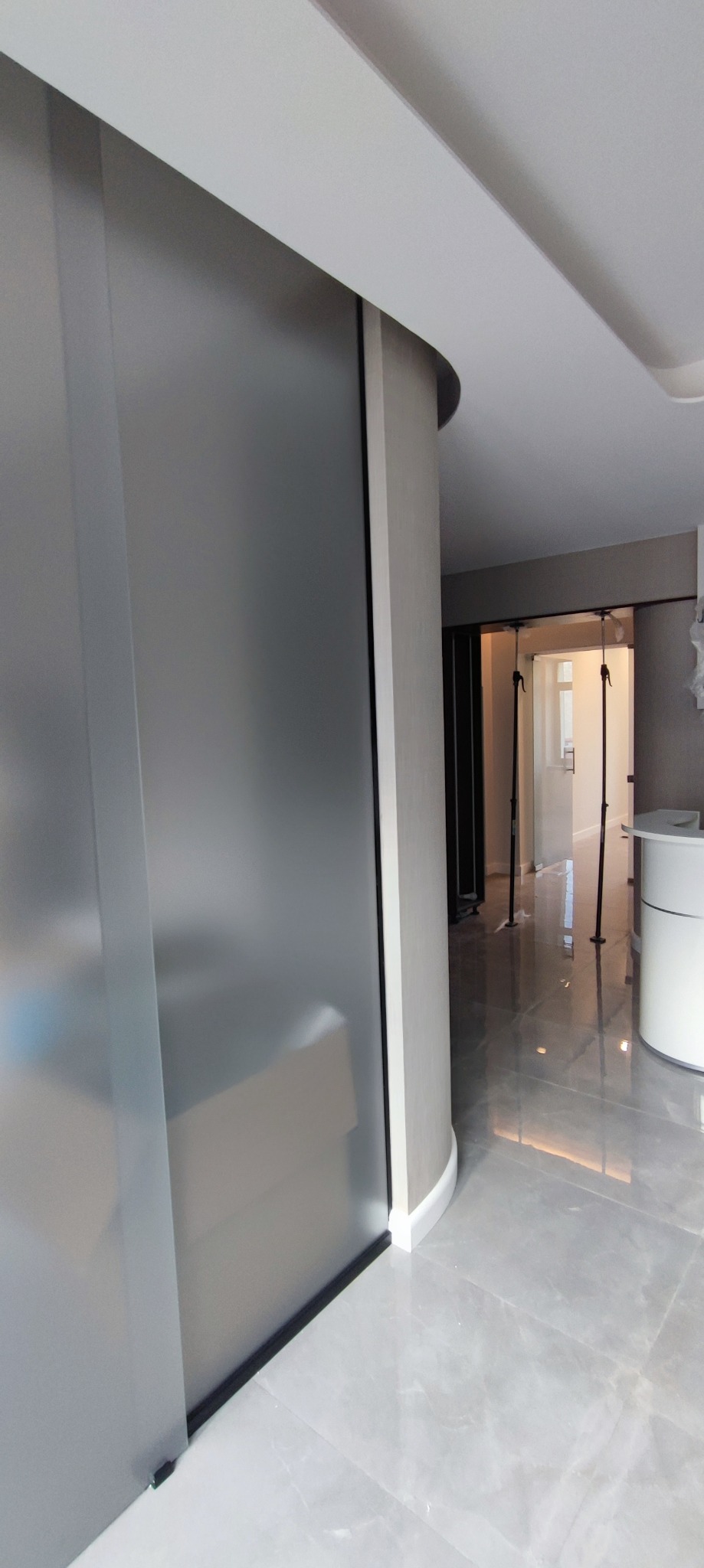 Nowoczesne wnętrze z przesuwnymi, matowymi drzwiami szklanymi, szara marmurowa podłoga i zaokrąglona ściana z białym wykończeniem przy podłodze.