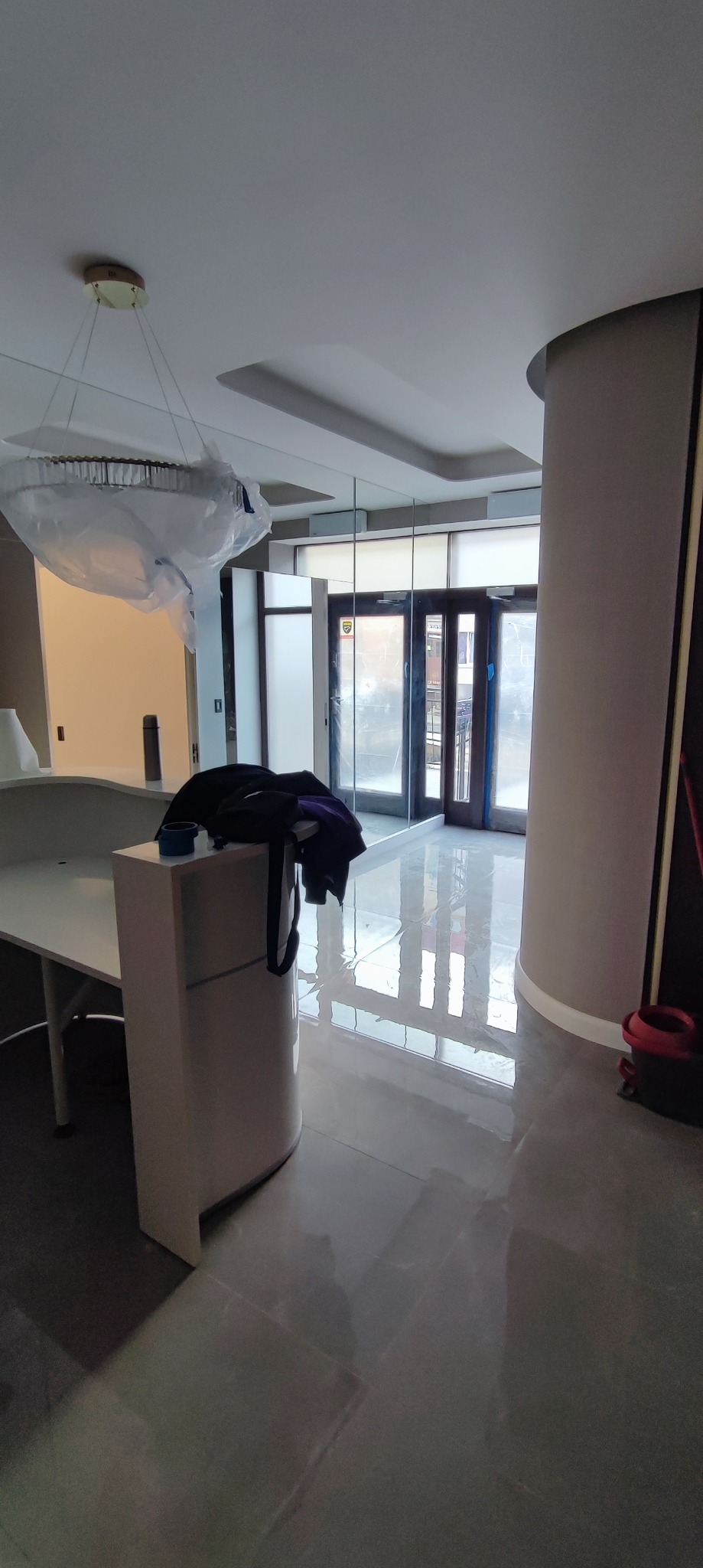 Świeżo położone, błyszczące płytki podłogowe w korytarzu, odbijające światło z okien i lampy pod sufitem, z widocznym fragmentem mebla i kolumny. Lampa sufitowa zabezpieczona folią ochronną.