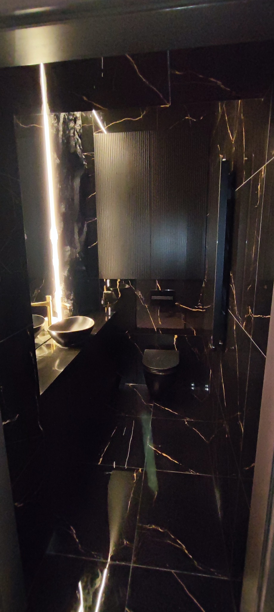 Elegancka łazienka wykończona czarnym marmurem z złotymi żyłami, z podświetlanym lustrem i umywalką nablatową, widok z perspektywy wejścia.