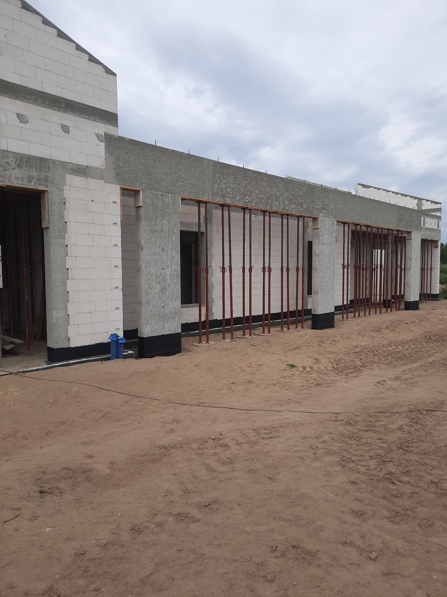 Wznoszenie ścian budynku z betonu komórkowego Ytong, widoczne stalowe stemple podtrzymujące konstrukcję, czarna izolacja przeciwwilgociowa fundamentów, ziemisty teren budowy.