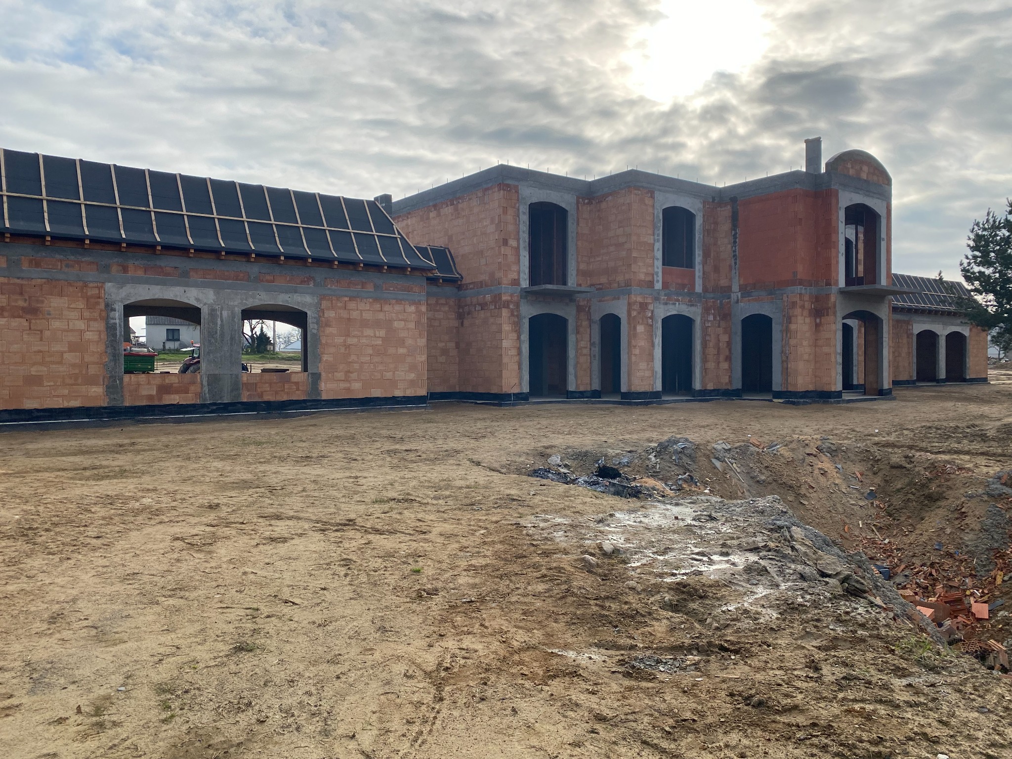 Surowy stan dwukondygnacyjnego domu z czerwonej cegły z widocznymi otworami okiennymi i częściowo wykonanym dachem na tle pochmurnego nieba, teren wokół w stanie przygotowania pod dalsze prace.