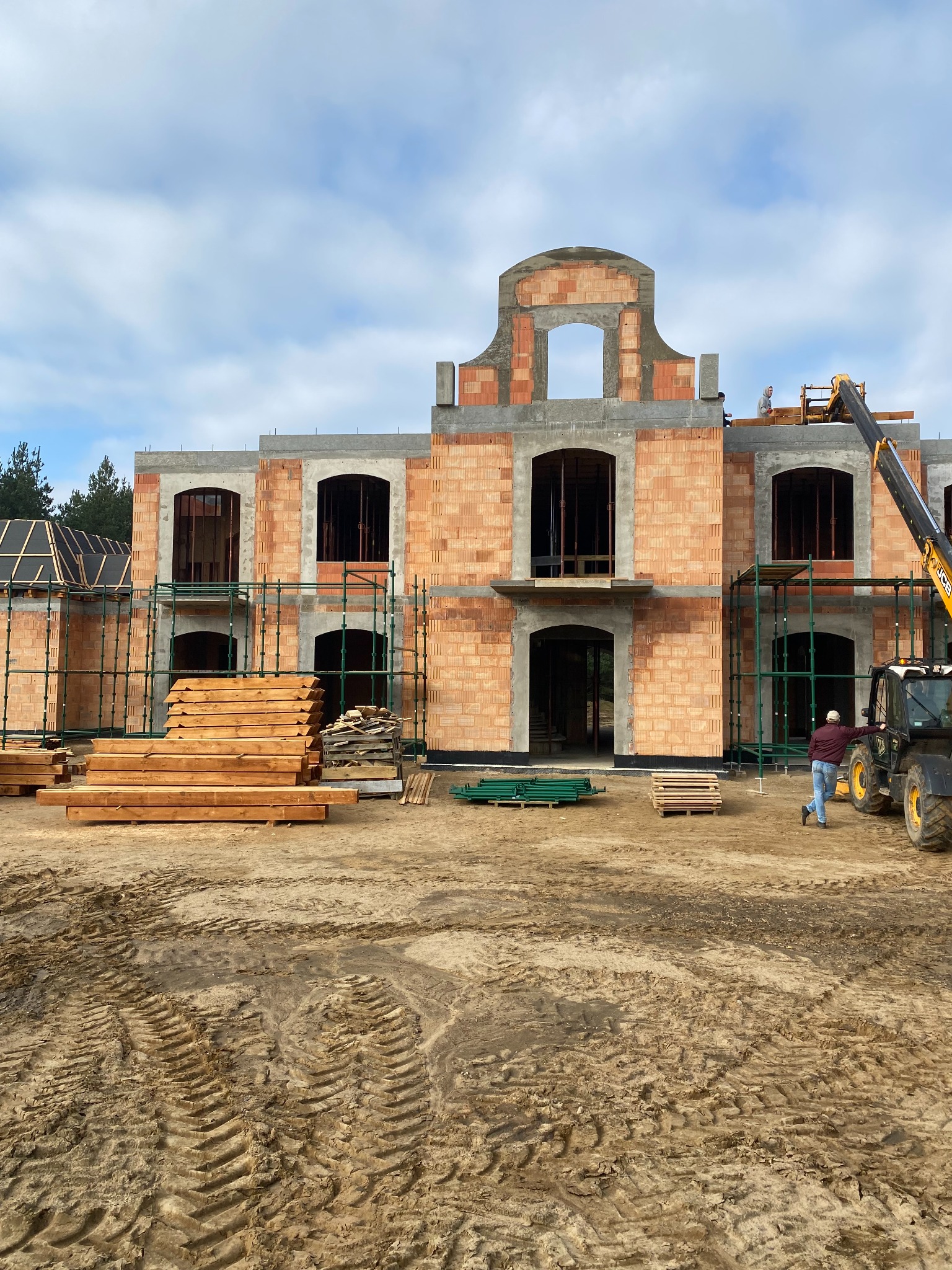 Wznoszenie dwukondygnacyjnego budynku z cegły, widoczne rusztowania, stos drewna i maszyna budowlana z operatorem w trakcie pracy.