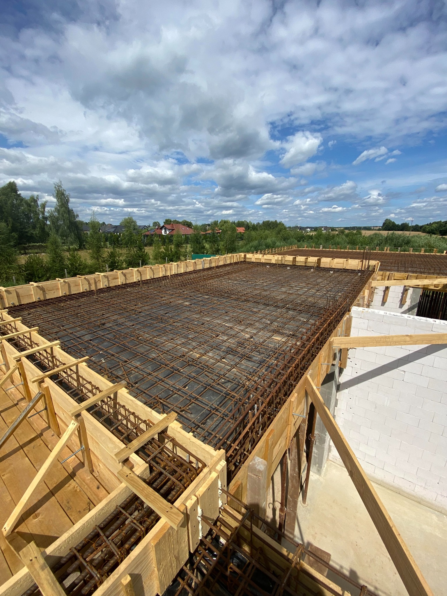 Zbrojenie płyty fundamentowej z szalunkiem drewnianym i widokiem na okoliczne domy i zachmurzone niebo.