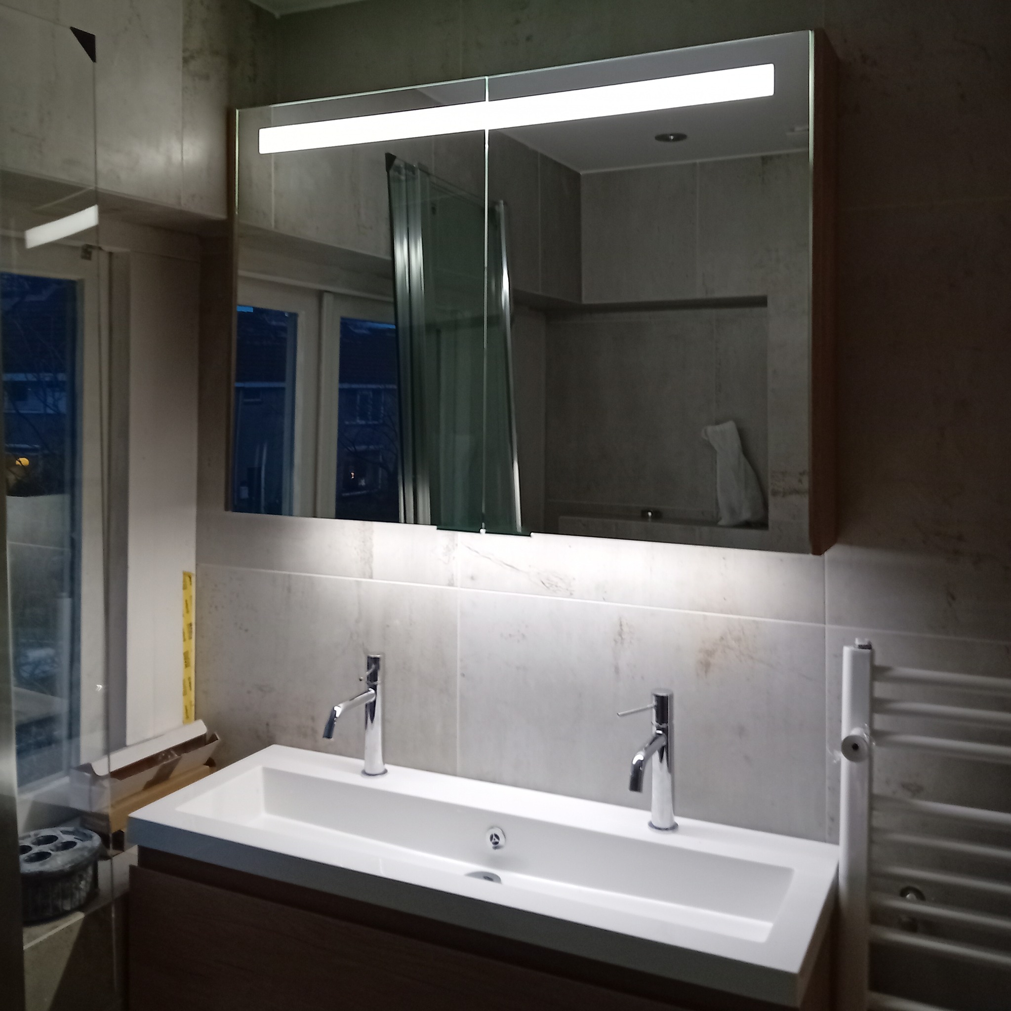 Nowoczesna łazienka z podwójną umywalką, lustrem z oświetleniem LED i grzejnikiem drabinkowym, widoczne okno z odbiciem budynku na zewnątrz.