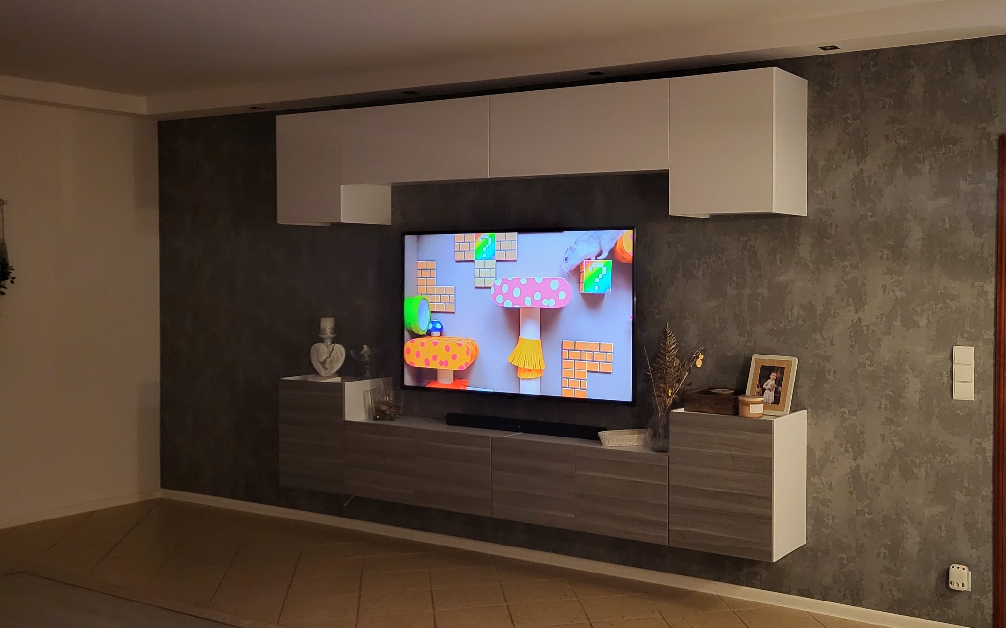 Nowoczesny salon z szarą ścianą o fakturze betonu, białe szafki wiszące i szafka RTV z frontami w odcieniach szarości, na szafce dekoracje i ramka ze zdjęciem, a na ścianie wisi telewizor.