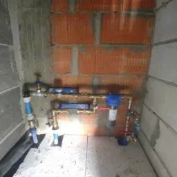 Fragment instalacji wodnej z filtrem i zaworami, zamontowany w narożniku pomieszczenia z surowymi ścianami z betonu kom&oacute;rkowego i cegły dziurawki.