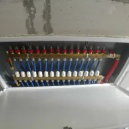 Rozdzielacz ogrzewania podłogowego w szafce z widocznymi zaworami, przepływomierzami i niebieskimi rurami, zamontowany przy surowej ścianie z betonu kom&oacute;rkowego.