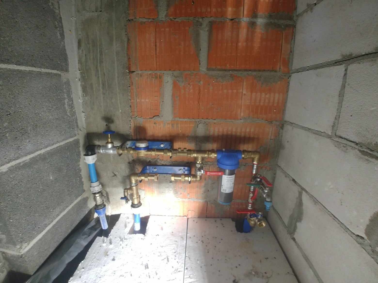 Fragment instalacji wodnej z filtrem i zaworami, zamontowany w narożniku pomieszczenia z surowymi ścianami z betonu komórkowego i cegły dziurawki.