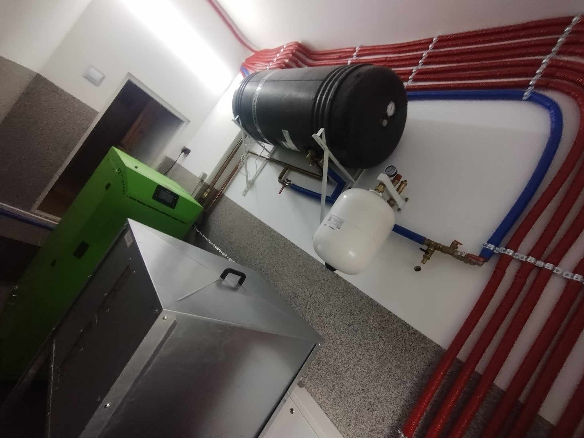 Kotłownia z zielonym kotłem, czarnym zbiornikiem akumulacyjnym, białym naczyniem przeponowym i rurami instalacji grzewczej w kolorze czerwonym i niebieskim, zamontowanymi na ścianie.