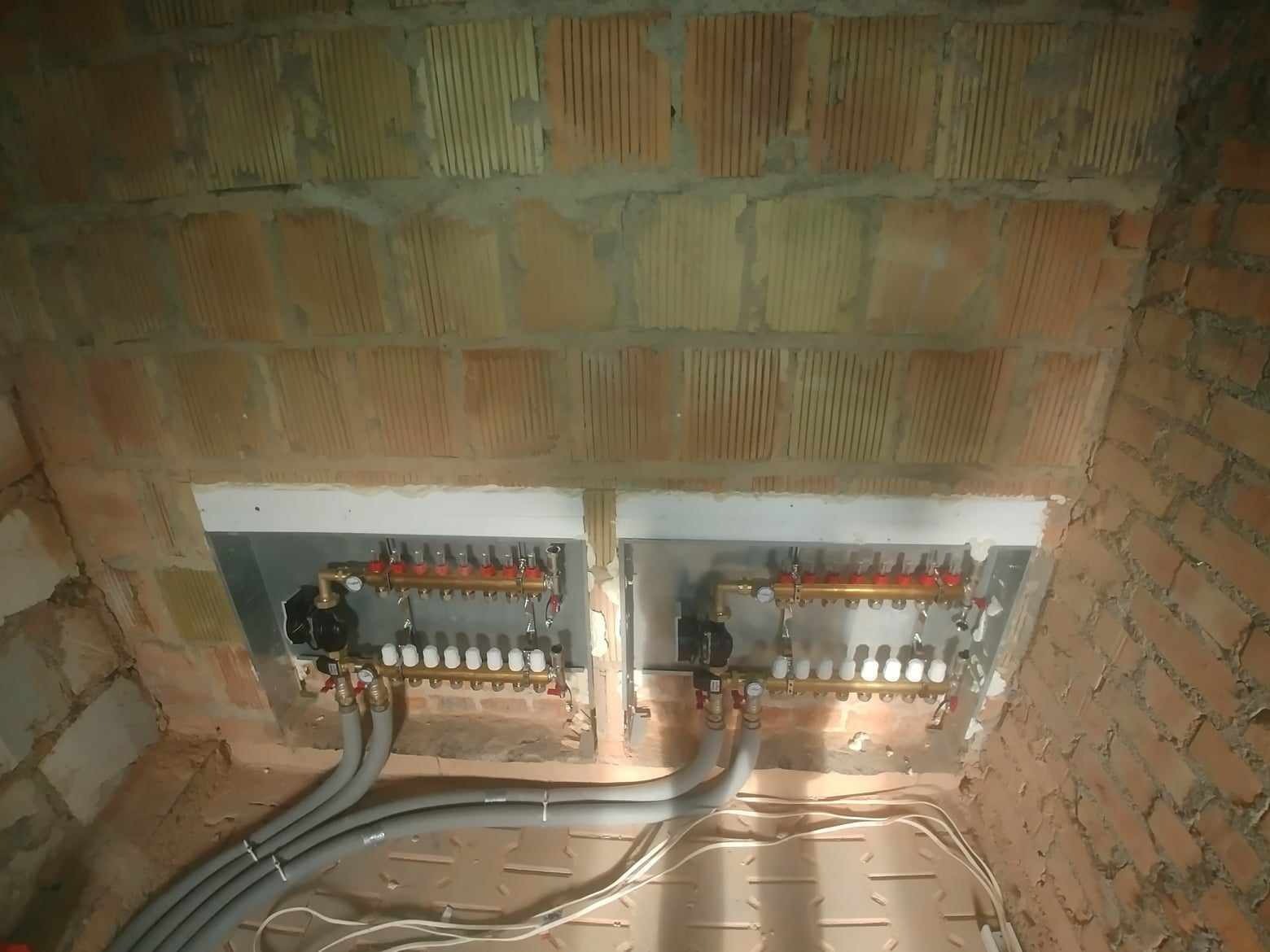 Rozdzielacze ogrzewania podłogowego zamontowane w ścianie z cegły dziurawki, widoczne szare rury doprowadzające wodę i białe zawory termostatyczne.