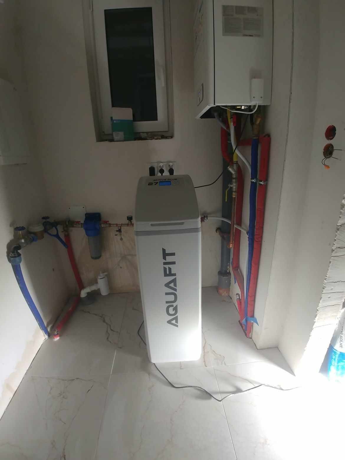 Szary, kompaktowy zmiękczacz wody Aquafit z podłączonymi rurami instalacji wodnej w pomieszczeniu z płytkami imitującymi marmur. Na ścianie widoczny bojler i okno.