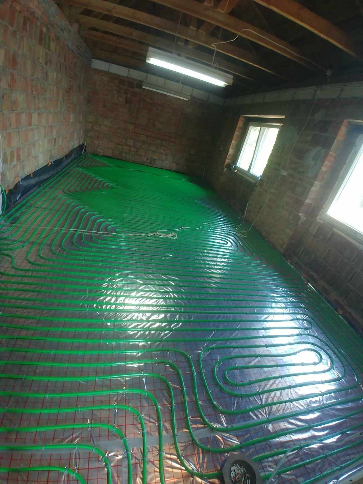 Zielone rury ogrzewania podłogowego ułożone na folii i siatce w surowym pomieszczeniu z ceglanymi ścianami i oknami dachowymi, przed wylaniem posadzki.