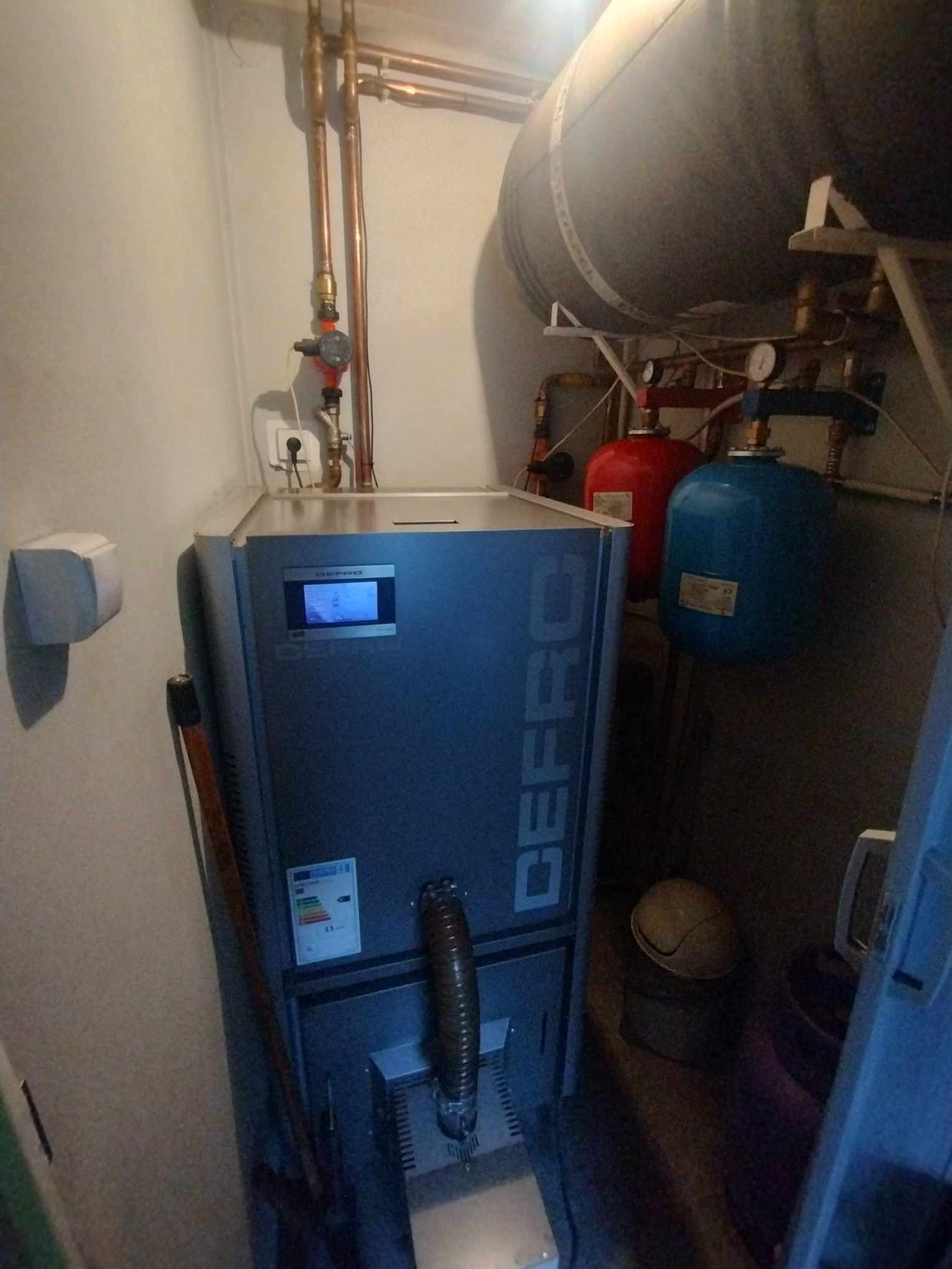 Kocioł grzewczy marki DEFRO w kotłowni, z widocznymi rurami miedzianymi, zbiornikiem akumulacyjnym i naczyniami przeponowymi.