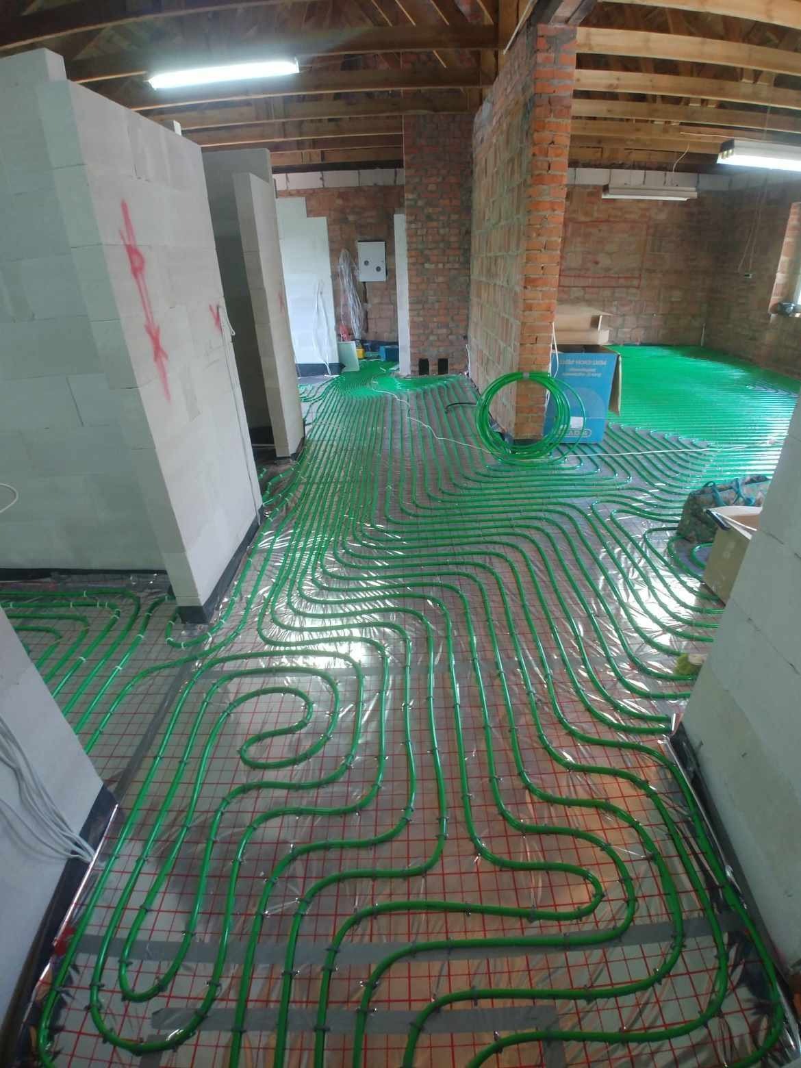 Wnętrze w trakcie prac instalacyjnych z widocznymi pętlami zielonych rur na metalowej siatce, przygotowanych do zalania w posadzce, ściany z cegły i bloczków betonowych w surowym stanie.