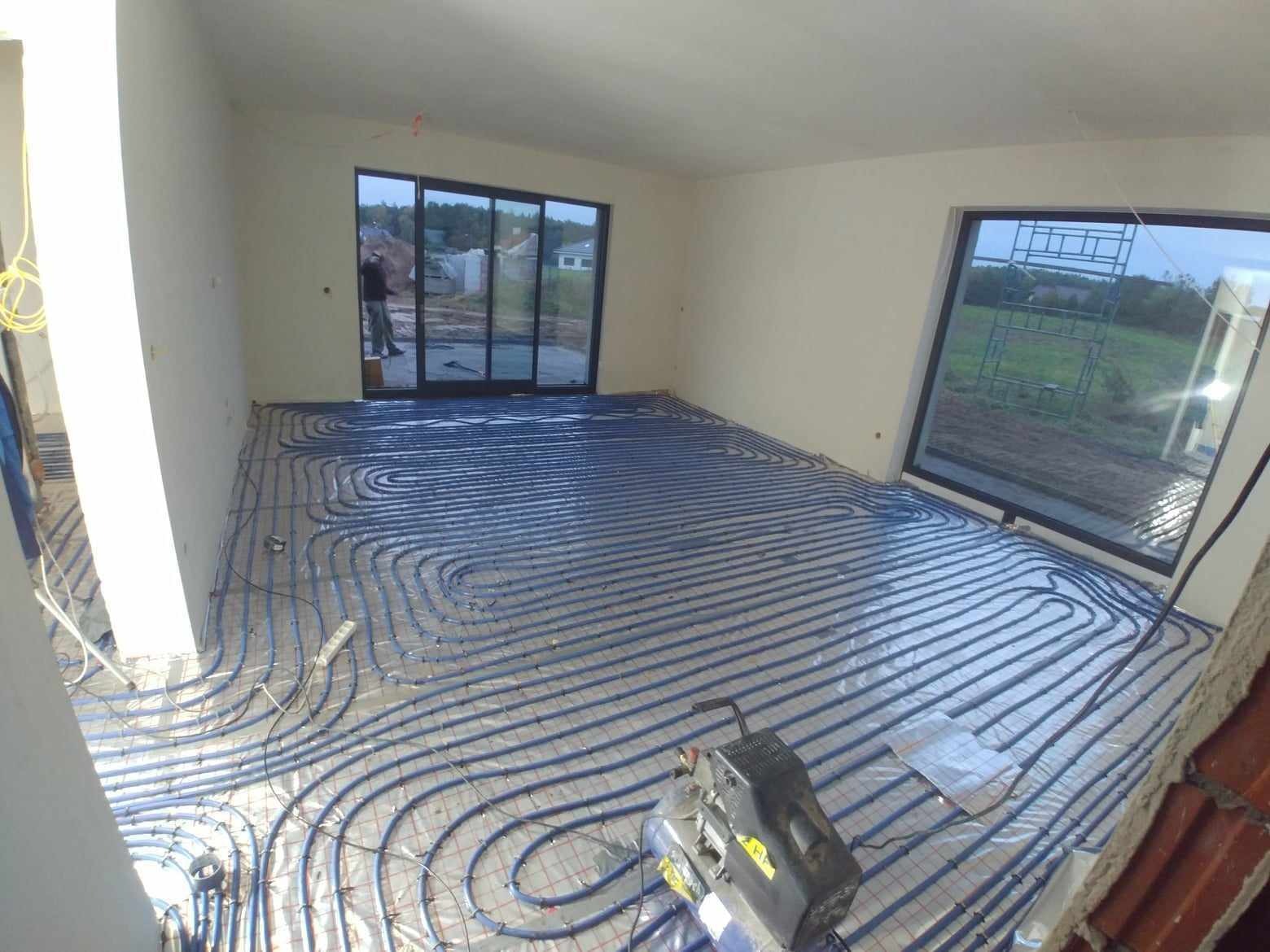 Niebieskie rury ogrzewania podłogowego ułożone spiralnie na folii izolacyjnej w surowym pomieszczeniu z dużymi oknami i drzwiami balkonowymi, widoczny kompresor.