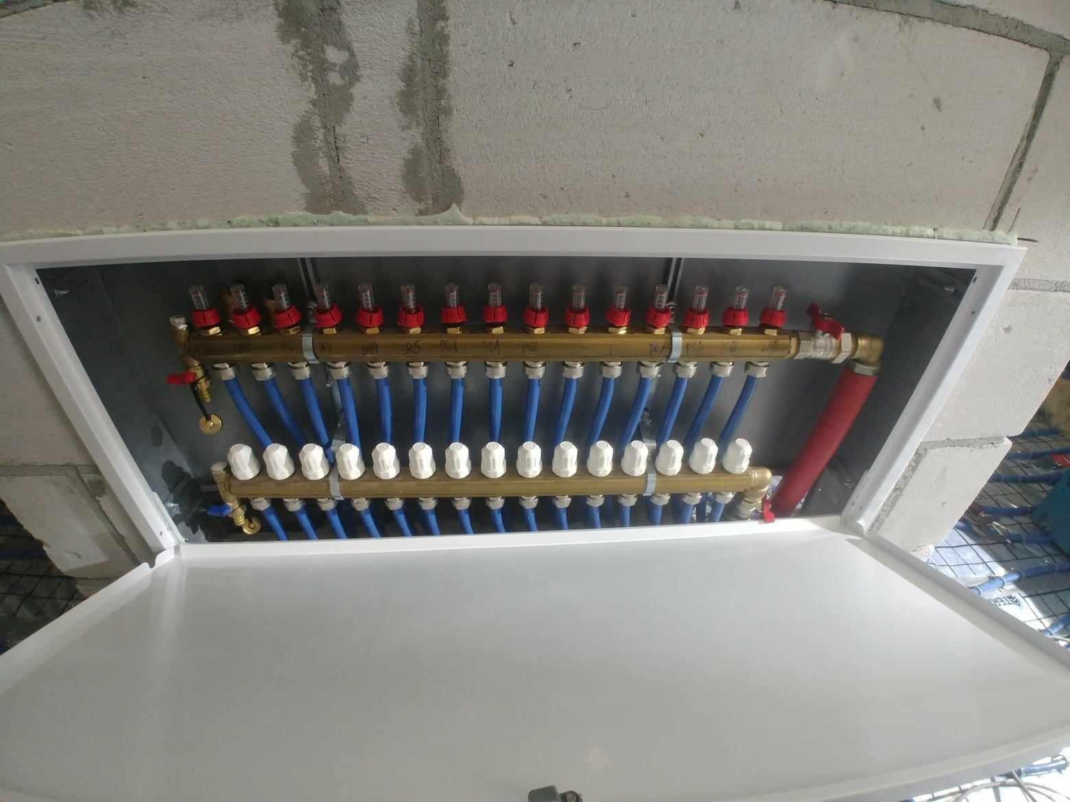 Rozdzielacz ogrzewania podłogowego w szafce z widocznymi zaworami, przepływomierzami i niebieskimi rurami, zamontowany przy surowej ścianie z betonu komórkowego.