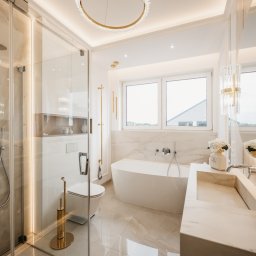 ALL DESIGN INSIDE Aleksandra Lepka - Elegancka łazienka w bieli i złocie: kabina prysznicowa ze szklanymi drzwiami, wolnostojąca wanna, umywalka z marmuru, okrągłe lustro z podświetleniem LED i dekoracyjny żyrandol.