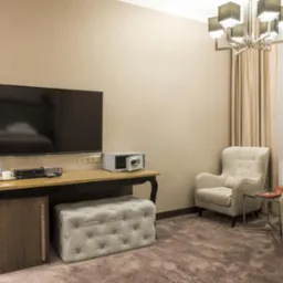 Pokój hotelowy w odcieniach beżu z telewizorem, minibarem, sejfem, pufą z guzikami i krzesłem z poduszką.