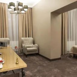 Pokój hotelowy z beżowymi zasłonami, wzorzystą tapetą i brązową wykładziną; na pierwszym planie stolik z czajnikiem, szklankami i filiżankami.