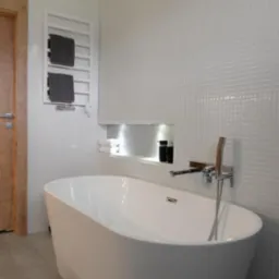 Nowoczesna, biała wanna wolnostojąca z chromowaną baterią, na tle ściany wyłożonej drobną, białą mozaiką i drewnianych drzwi.