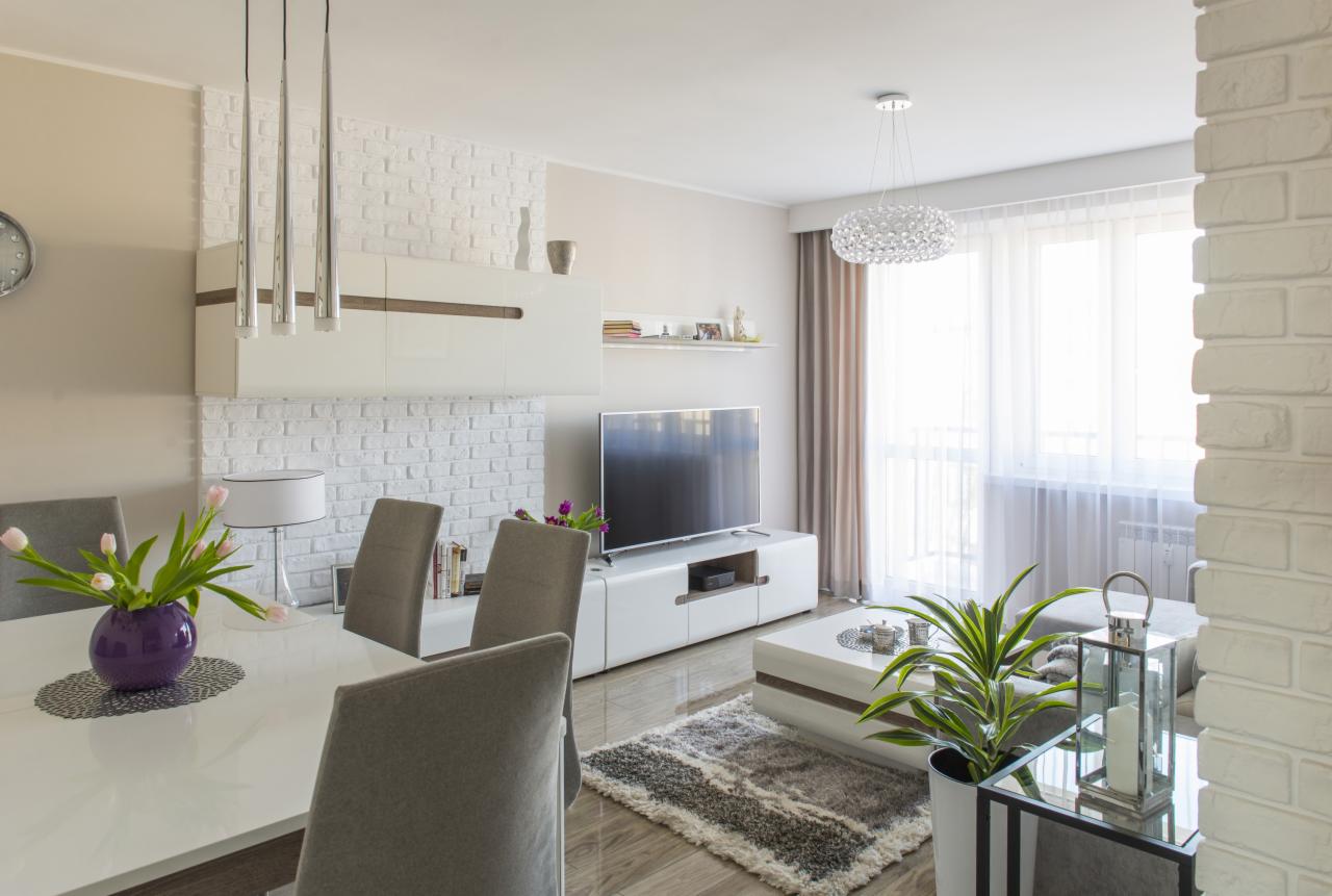 Jasne, nowoczesne wnętrze z białymi ceglanymi ścianami, drewnianą podłogą, białymi meblami i szarymi krzesłami. Na stole fioletowy wazon z różowymi tulipanami.