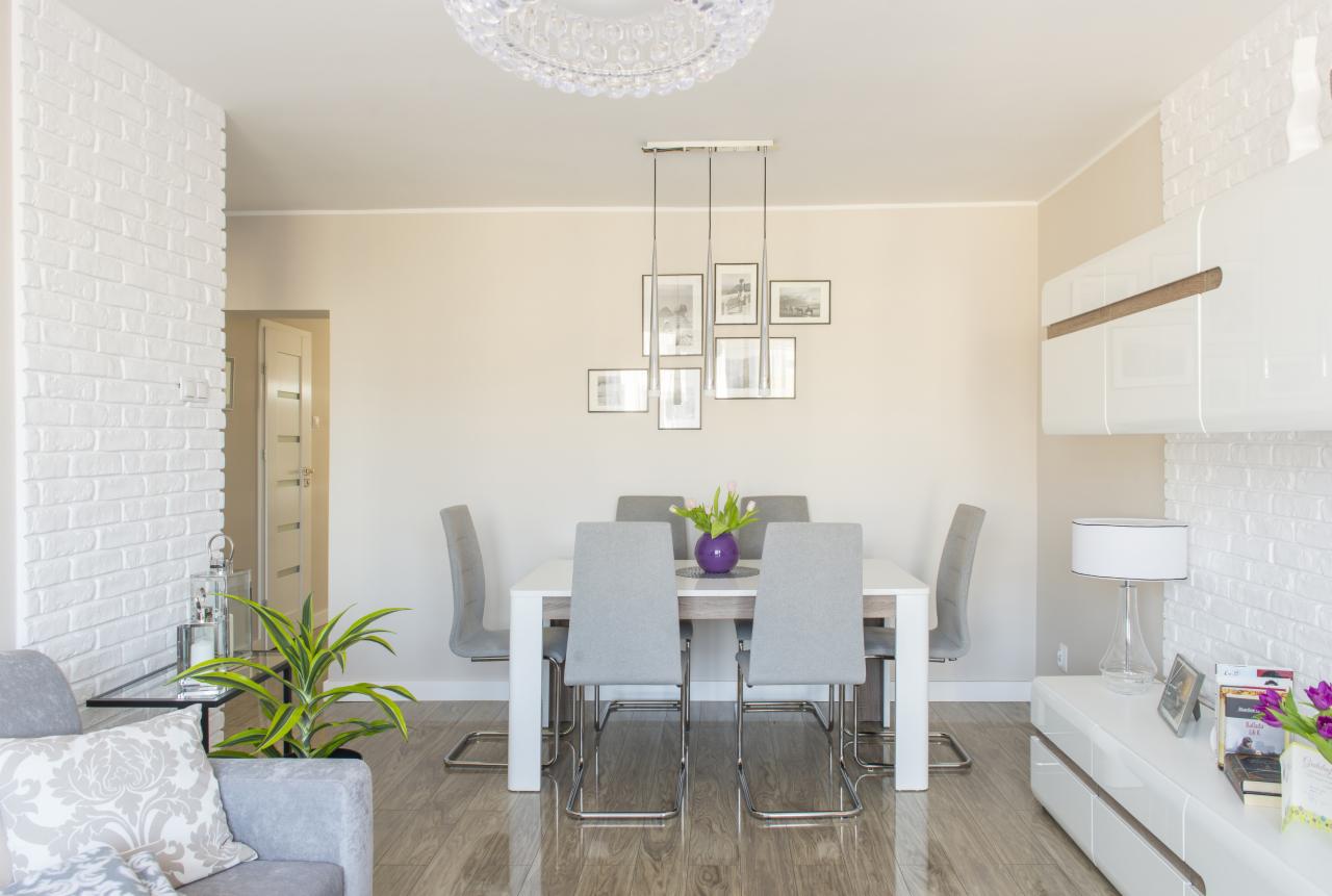 Jasne wnętrze jadalni z białym stołem, szarymi krzesłami, fioletowym wazonem z tulipanami, oświetleniem wiszącym z elementami zdjęć, oraz ścianami z białej cegły i drewnopodobną podłogą.