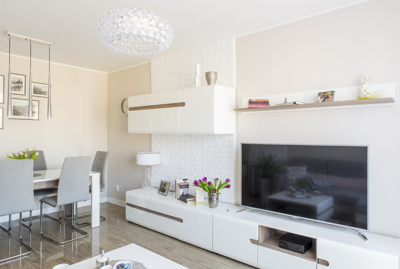 Jasne wnętrze salonu z ceglaną ścianą, białymi meblami, szarymi krzesłami przy stole i dekoracyjnym oświetleniem sufitowym