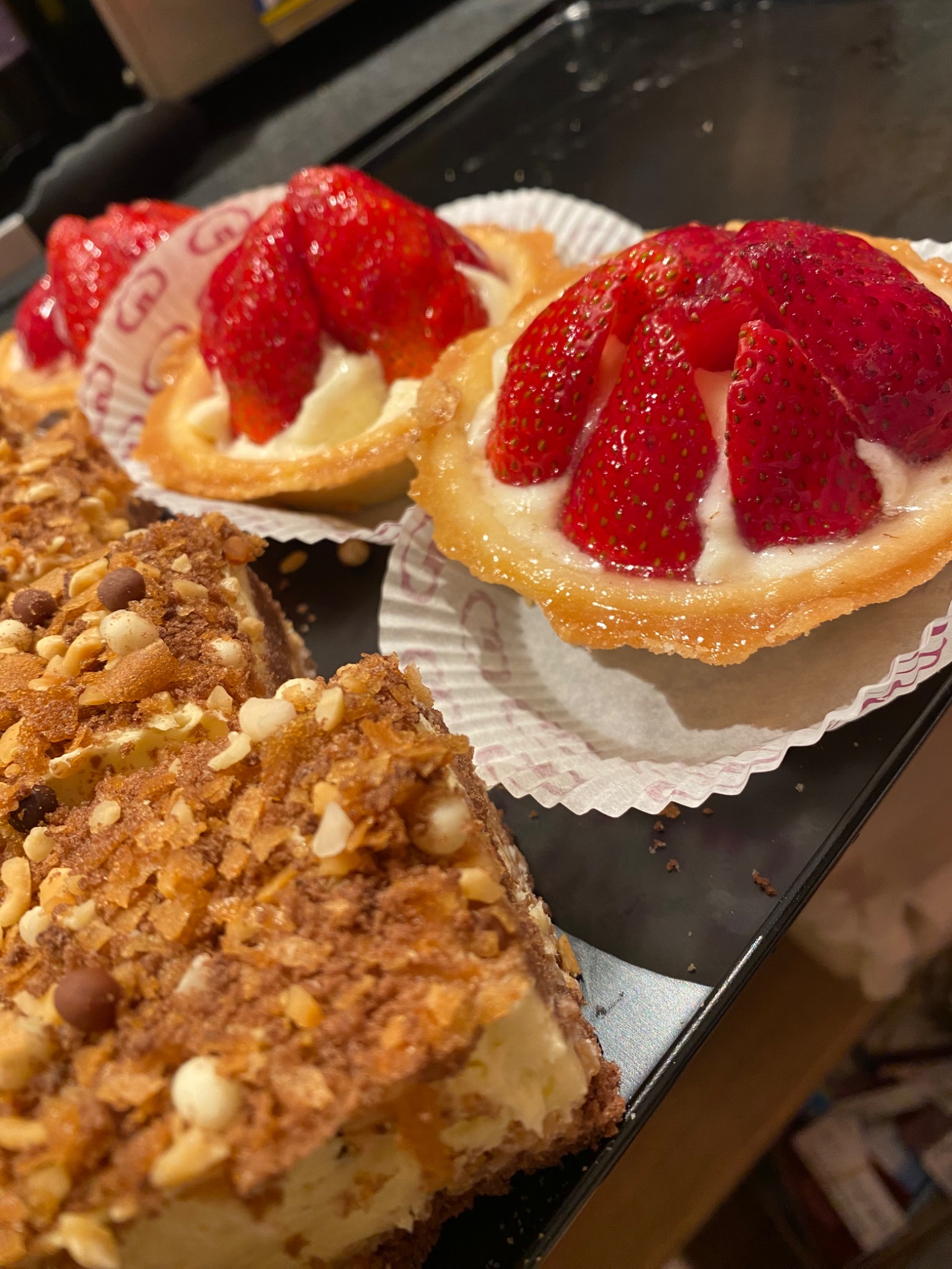 Tarty z kremem i świeżymi truskawkami w papierowych foremkach obok ciasta orzechowego z kremem na czarnej tacy.