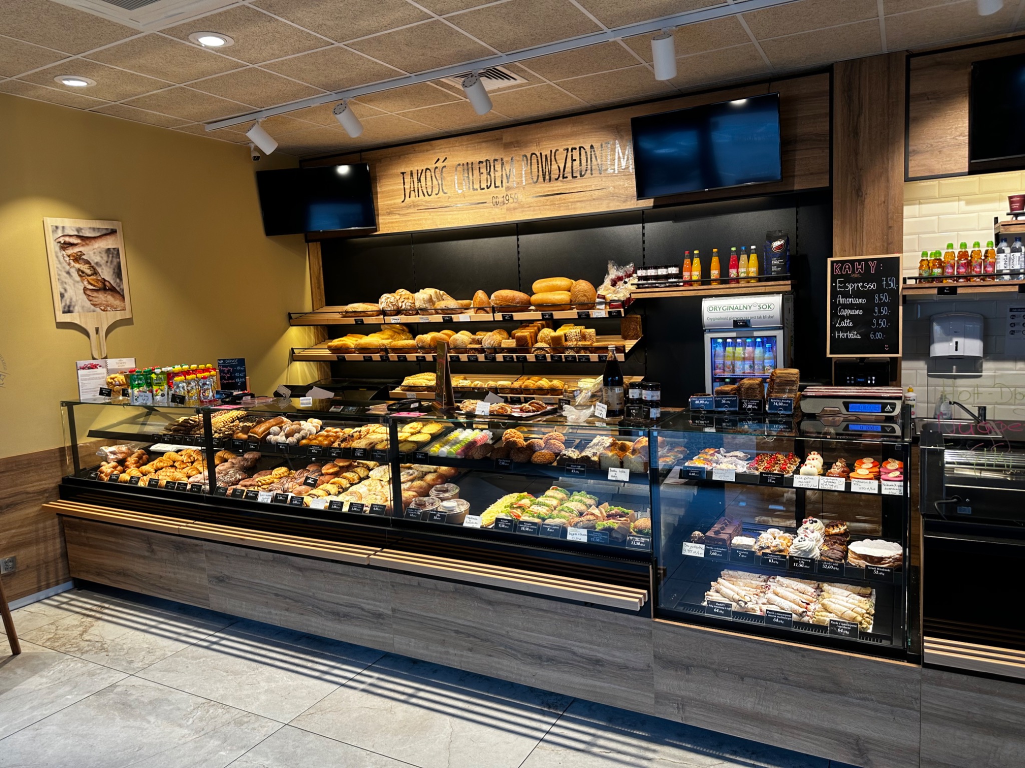 Wnętrze cukierni z pełnymi półkami pieczywa i słodkości w przeszklonych witrynach, tablica z cenami kaw i logo 'Jakość chlebem powszednim od 1959'.
