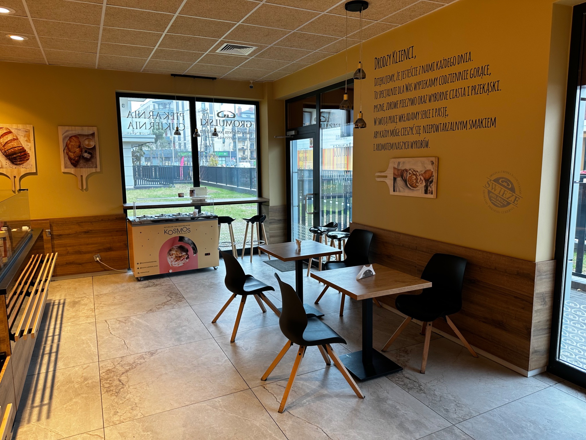 Wnętrze kawiarni z jasnymi ścianami, stolikami z czarnymi krzesłami, dekoracyjnym napisem na ścianie i lodówką z logo firmy KOSMOS