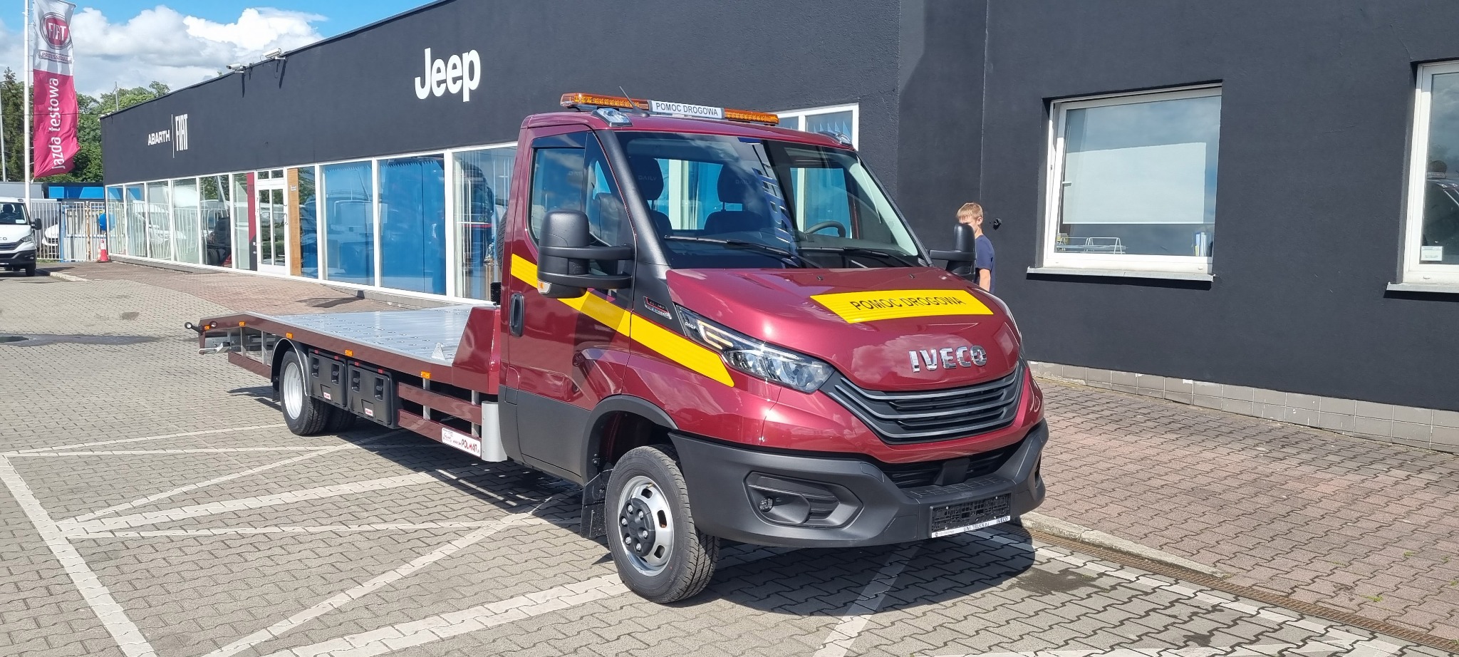 Czerwony samochód pomocy drogowej marki Iveco z żółtymi pasami i napisem 'Pomoc Drogowa' na tle salonu samochodowego Jeep i Abarth.