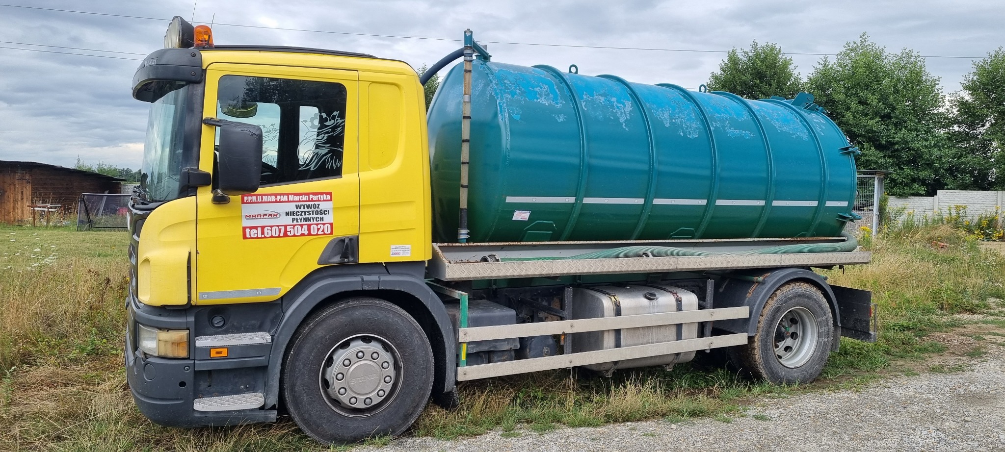 Żółta ciężarówka z zielonym zbiornikiem do wywozu nieczystości płynnych, z widocznym numerem telefonu i nazwą firmy na kabinie, stojąca na żwirowej drodze w otoczeniu zieleni.