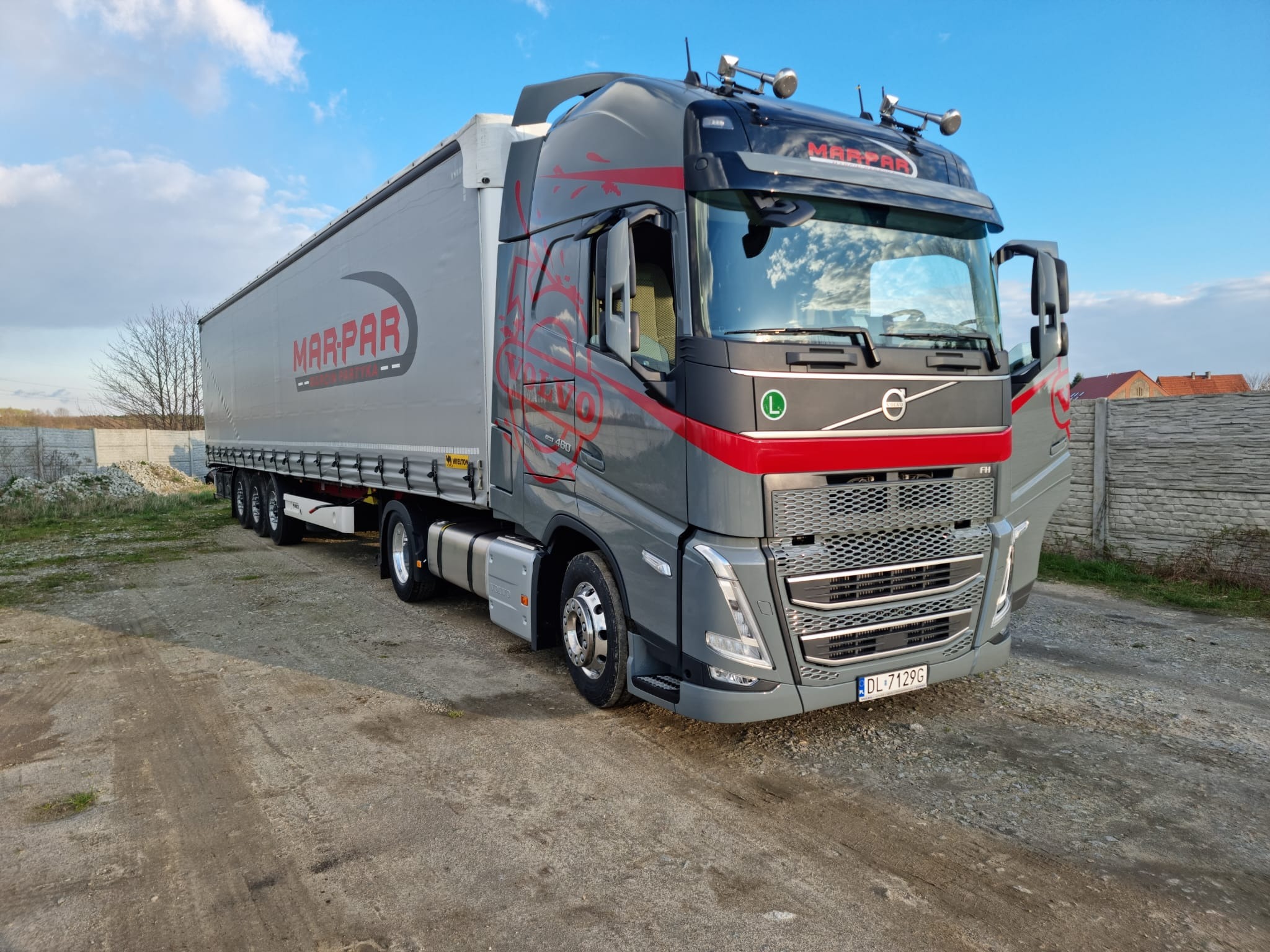 Szara ciężarówka Volvo z naczepą z logo Mar-Par zaparkowana na żwirowym podłożu, widoczna otwarta kabina kierowcy, błękitne niebo w tle.