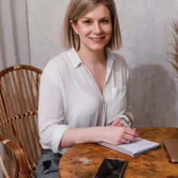 Uśmiechnięta blondynka w białej koszuli siedzi przy okrągłym, drewnianym stole, pisząc w notatniku; obok leży smartfon i częściowo widoczny laptop.