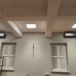 IS COOL Ireneusz Stasik - Dwa czarne klimatyzatory ścienne zamontowane symetrycznie nad oknami z roletami w jasnym pomieszczeniu z białymi grzejnikami i drabinką. Minimalistyczny design.