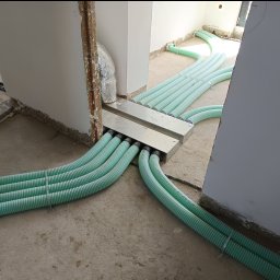 IS COOL Ireneusz Stasik - Rozprowadzenie instalacji wentylacyjnej z zielonych, karbowanych rur w surowym wnętrzu. Widoczne metalowe elementy mocujące i fragmenty ścian w trakcie wykończenia.