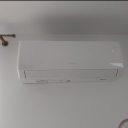 IS COOL Ireneusz Stasik - Biała klimatyzacja Gree zamontowana na ścianie w Dobczycach. Widoczny fragment karnisza i zasłony. Minimalistyczny design, czyste linie, oświetlenie LED sygnalizuje pracę urządzenia.