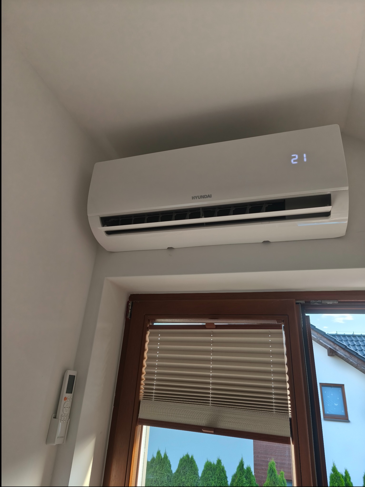 Biały klimatyzator Hyundai zamontowany nad oknem z brązową ramą i żaluzjami. Na ścianie obok wisi pilot. Widoczny fragment domu za oknem.