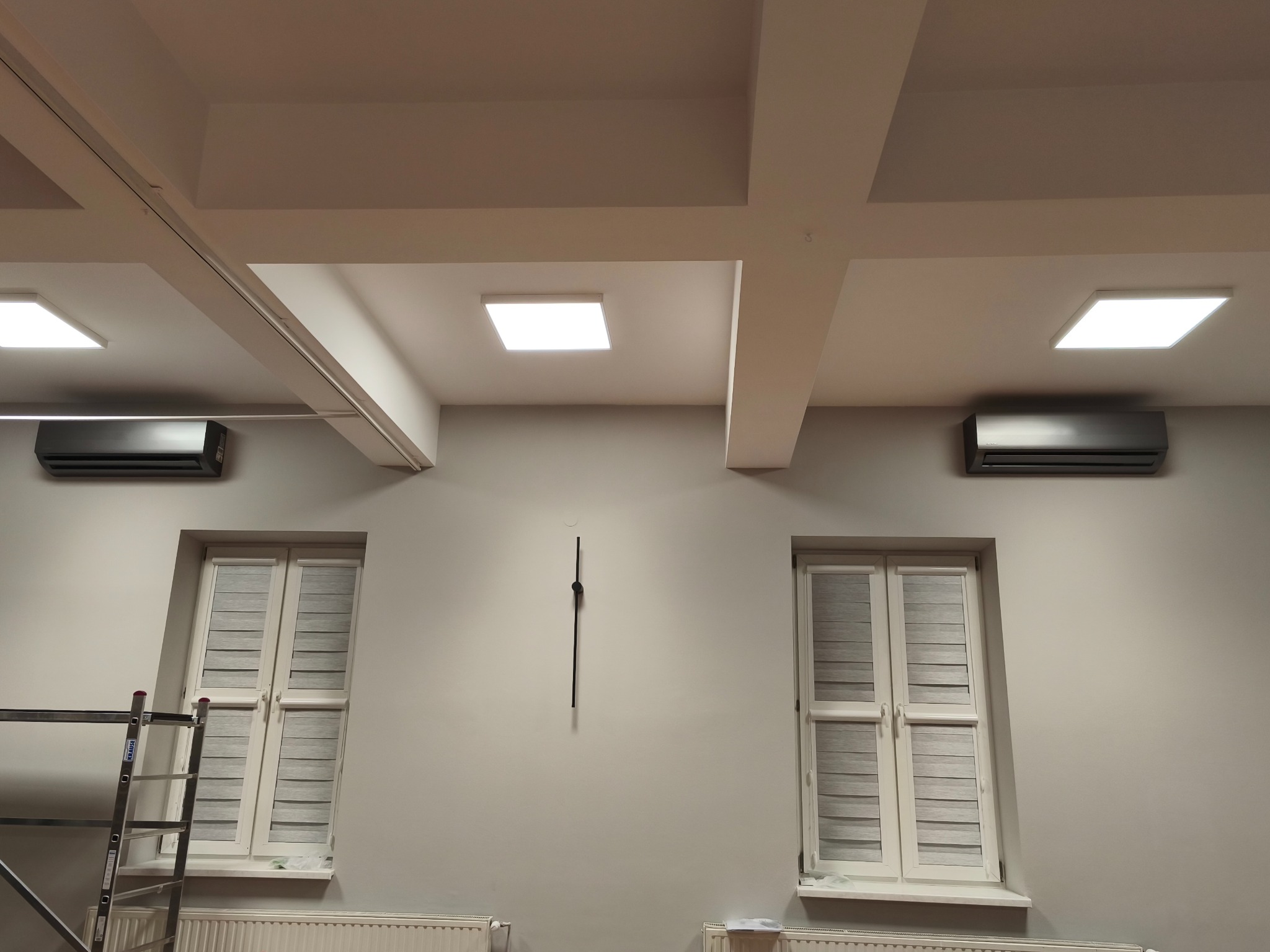 Dwa czarne klimatyzatory ścienne zamontowane symetrycznie nad oknami z roletami w jasnym pomieszczeniu z białymi grzejnikami i drabinką. Minimalistyczny design.