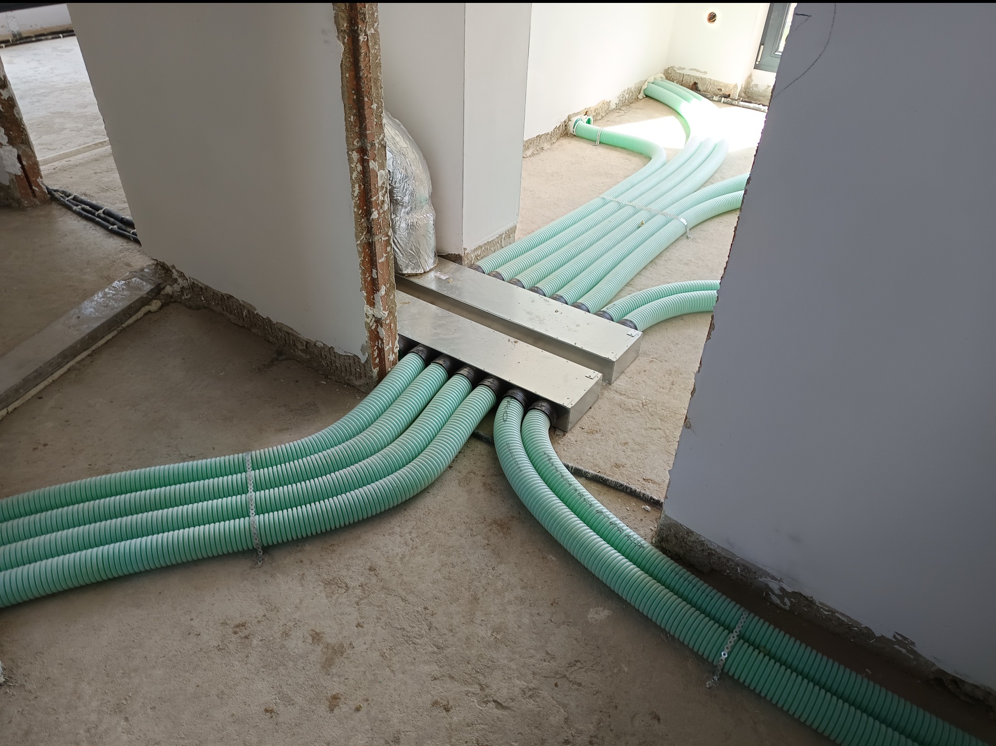 Rozprowadzenie instalacji wentylacyjnej z zielonych, karbowanych rur w surowym wnętrzu. Widoczne metalowe elementy mocujące i fragmenty ścian w trakcie wykończenia.