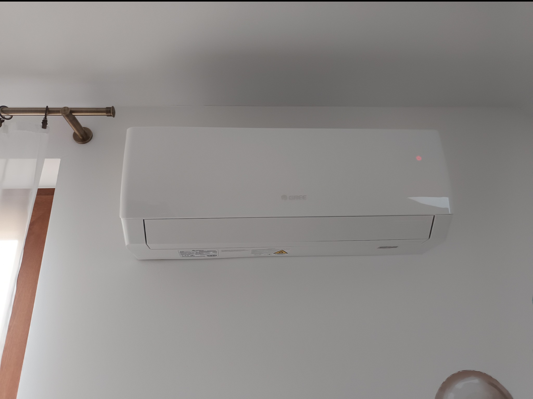 Biała klimatyzacja Gree zamontowana na ścianie w Dobczycach. Widoczny fragment karnisza i zasłony. Minimalistyczny design, czyste linie, oświetlenie LED sygnalizuje pracę urządzenia.