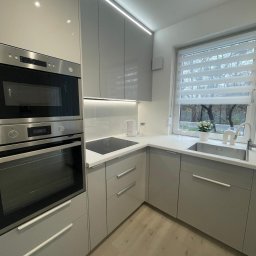 Pavel Kanenka - Jasna, nowoczesna kuchnia w odcieniach szarości z oknem, zabudową meblową i sprzętem AGD. Minimalistyczny design, oświetlenie LED i drewniana podłoga.