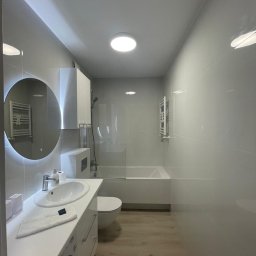 Pavel Kanenka - Nowoczesna łazienka w odcieniach szarości i bieli z okrągłym lustrem z podświetleniem LED, drewnopodobną podłogą i minimalistyczną armaturą.