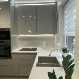 Pavel Kanenka - Nowoczesna kuchnia w szarościach z białym blatem, oświetleniem LED podszafkowym i minimalistyczną armaturą. Roślina doniczkowa dodaje świeżości.