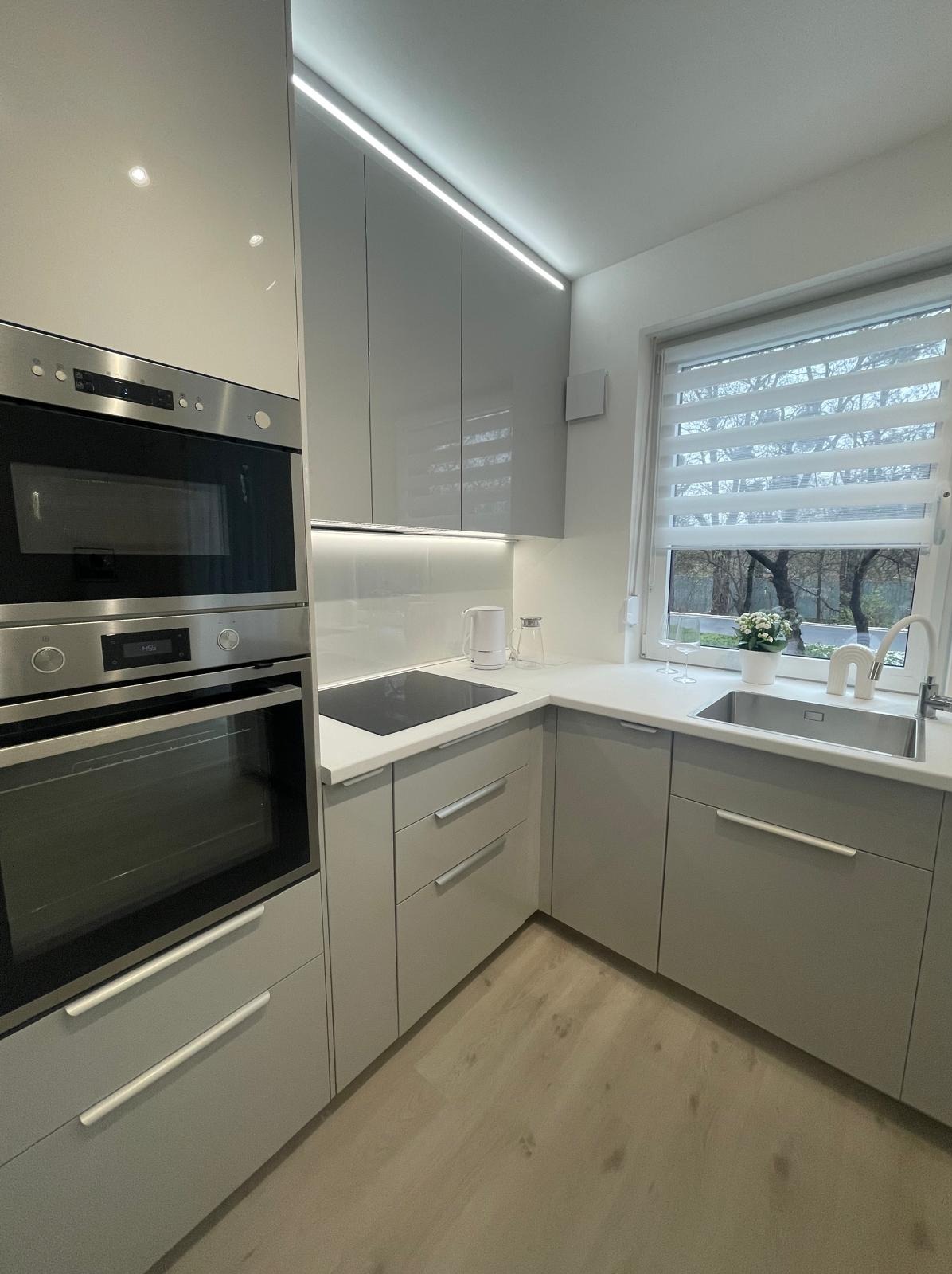 Jasna, nowoczesna kuchnia w odcieniach szarości z oknem, zabudową meblową i sprzętem AGD. Minimalistyczny design, oświetlenie LED i drewniana podłoga.