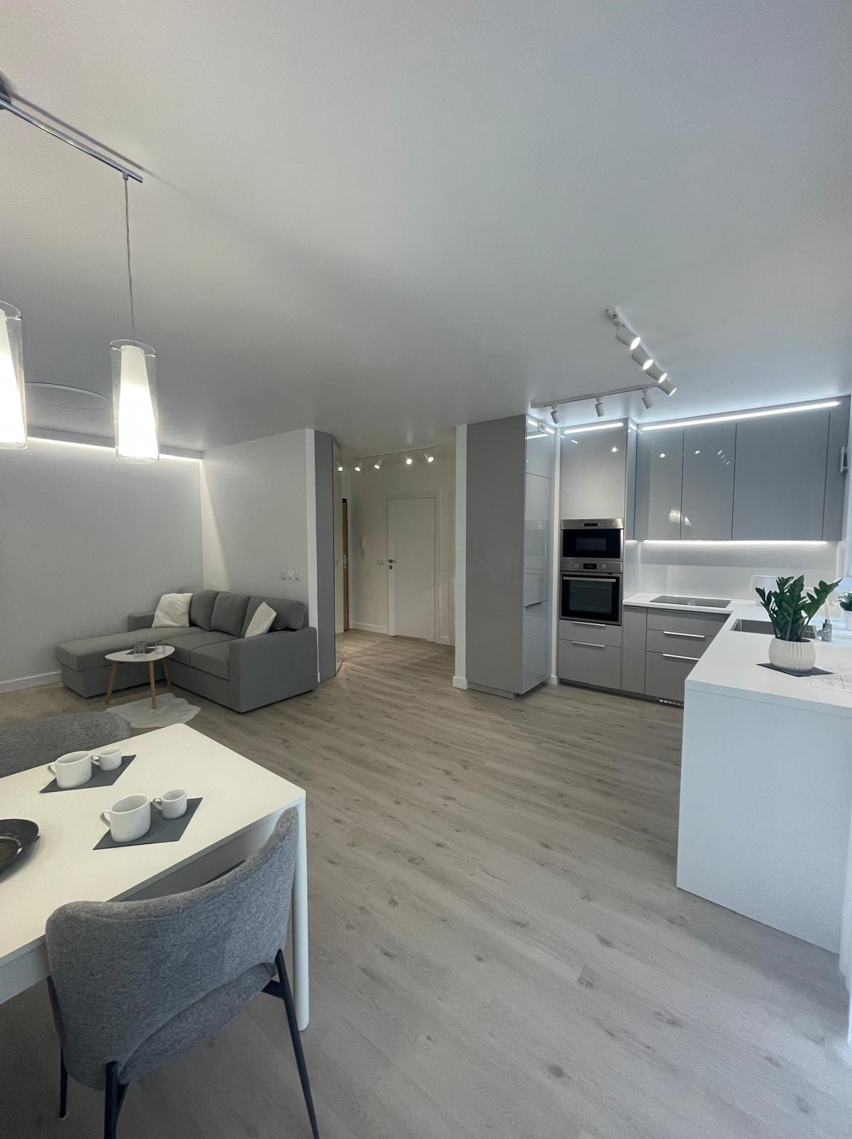 Nowoczesne wnętrze: szara kuchnia z wyspą, drewnopodobna podłoga, szara sofa w salonie. Minimalistyczny design, oświetlenie LED. Widok z perspektywy jadalni.