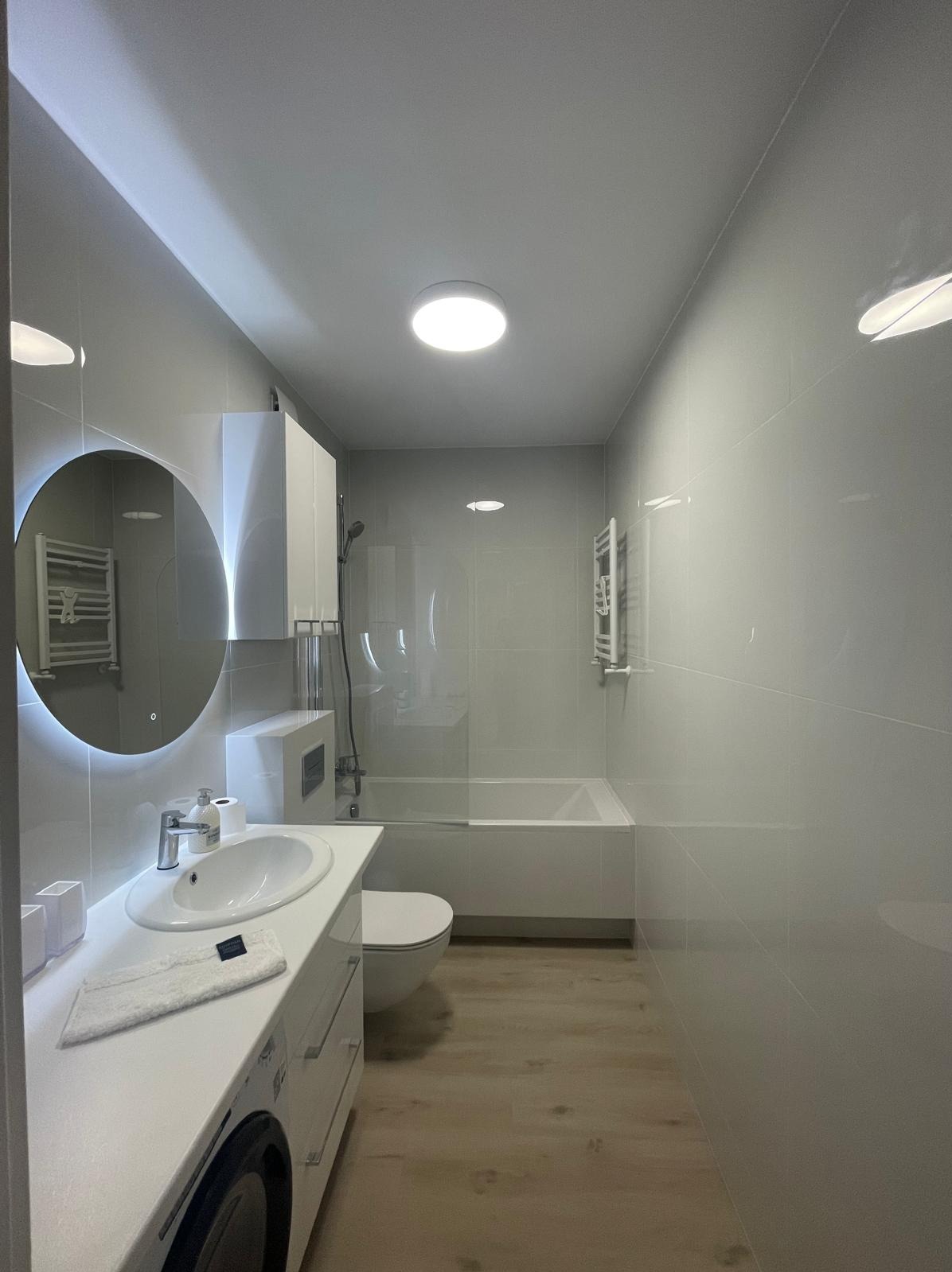 Nowoczesna łazienka w odcieniach szarości i bieli z okrągłym lustrem z podświetleniem LED, drewnopodobną podłogą i minimalistyczną armaturą.