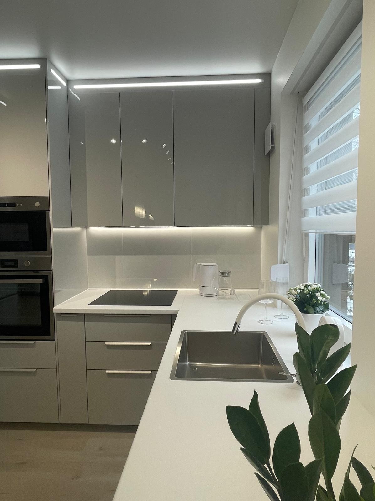 Nowoczesna kuchnia w szarościach z białym blatem, oświetleniem LED podszafkowym i minimalistyczną armaturą. Roślina doniczkowa dodaje świeżości.