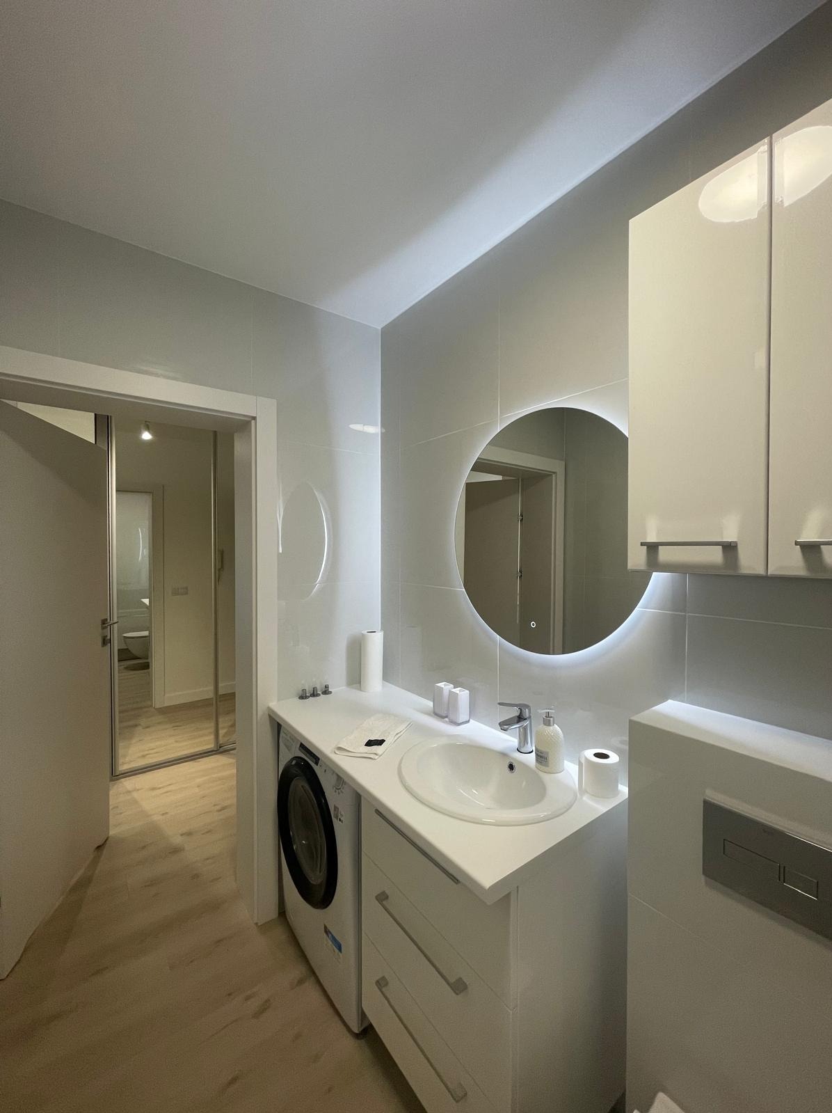 Nowoczesna łazienka w odcieniach szarości z okrągłym lustrem z podświetleniem LED, białą umywalką i pralką pod blatem. Widoczne płytki na ścianach i drewnopodobna podłoga.